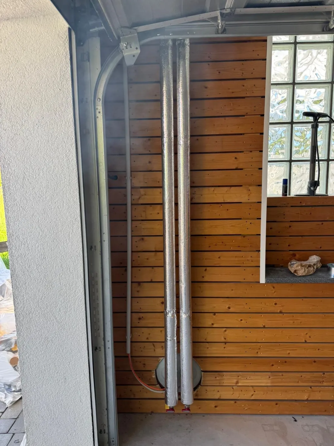 Zwei silbern gedämmte Leitungen kommen durch eine Kernbohrung und laufen an der Holzwand nach oben